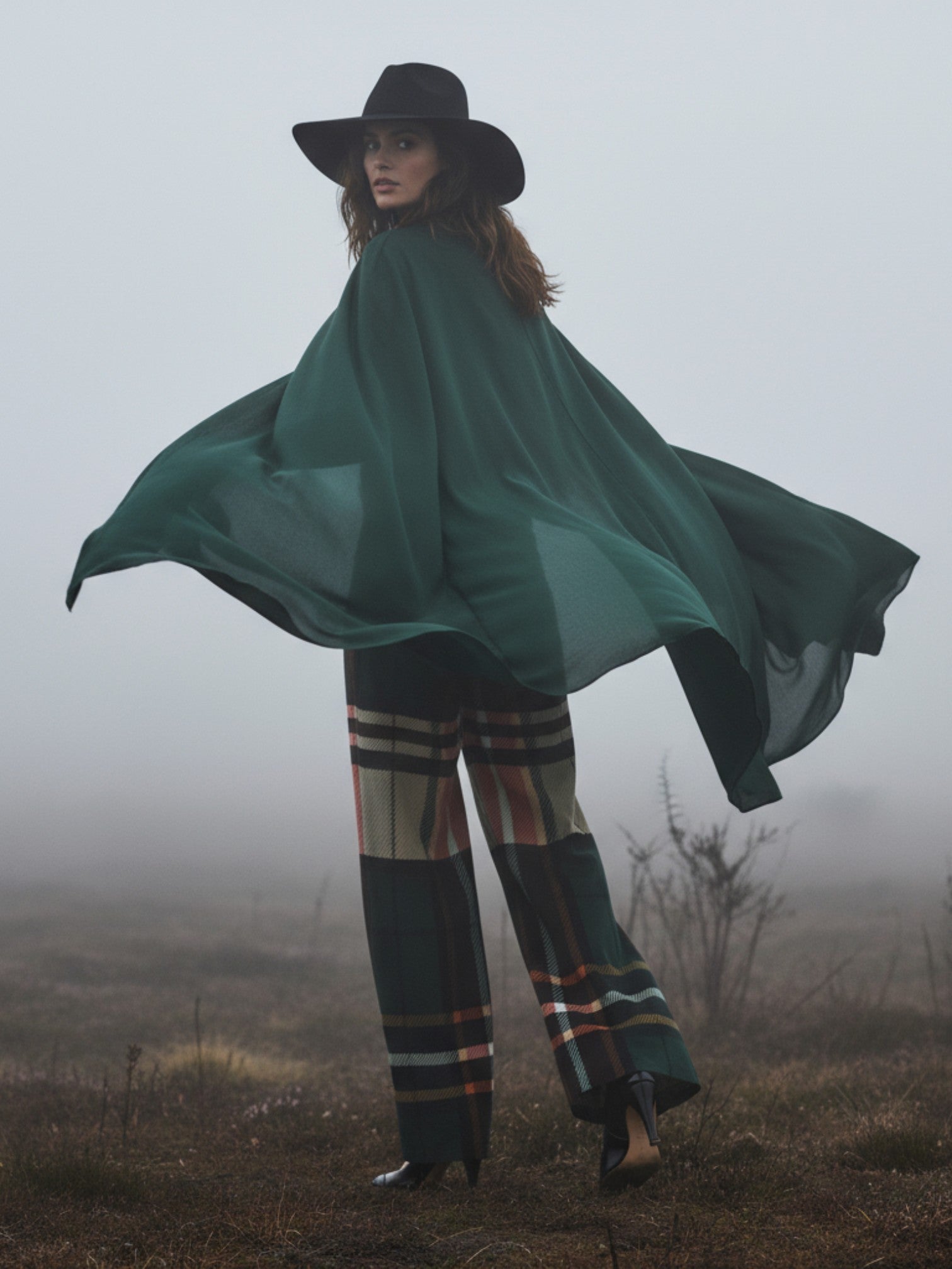 Woman wearing women's tartan wide-leg trousers with a flowing green cape and black hat in foggy outdoor setting